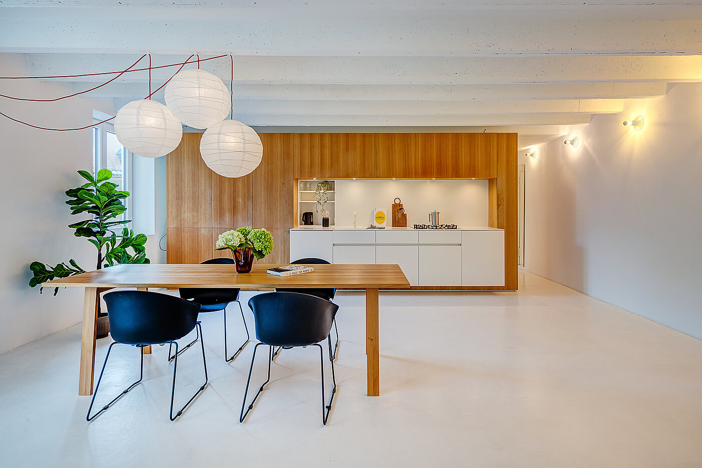 Home Staging einer Wohnung: Essbereich mit Holztisch, dunkelblauen Stühlen und Papierlampen vor offener Küche mit Holzfronten