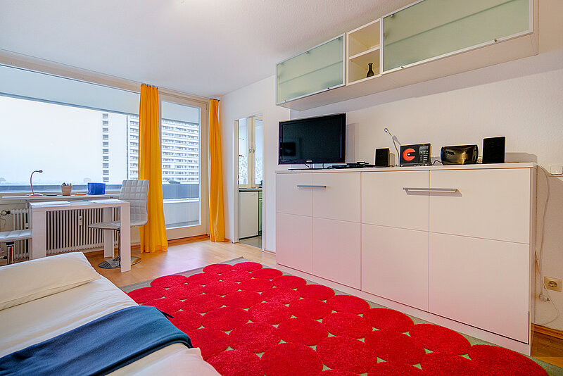 Apartment mit alter Möblierung Zu sehen ist ein Einzelbett links, gelbe Vorhänge, ein roter Teppich und ein weißes Sideboard mit Flachbildfernseher.