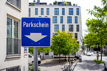 Zu sehen ist ein Straßenschild mit der Aufschrift Parkschein, im Hintergrund sind Neubauten