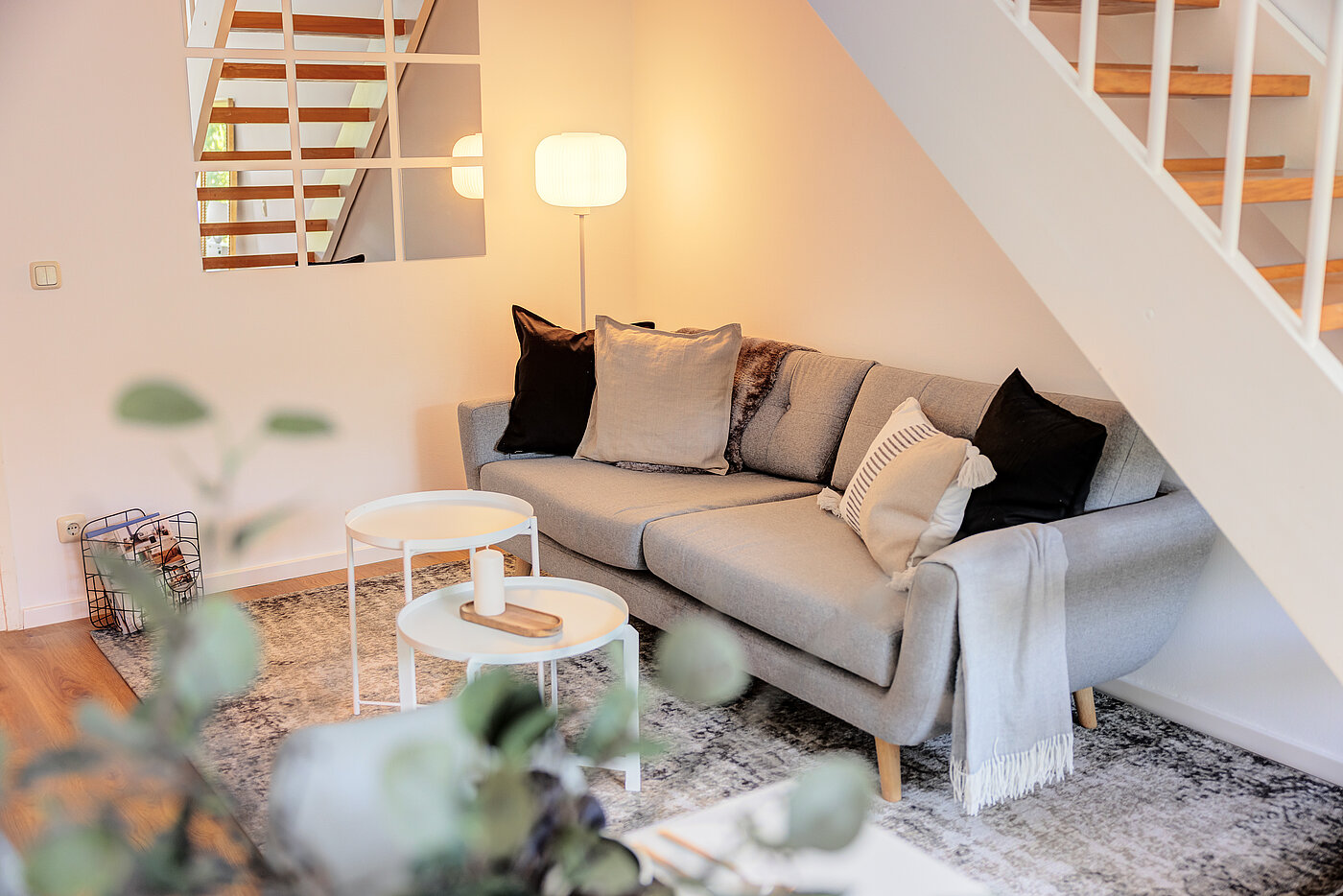Home Staging: gemütliche Sitzecke unter der Treppe mit grauem Sofa, Kissen, Stehlampe und runden Beistelltischen