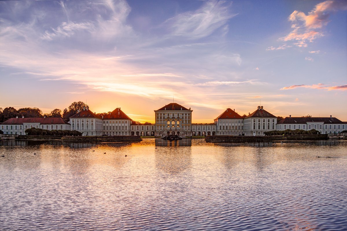 Zu sehen ist das Schloss Nmyphenburg bei Sonnenuntergang, im Vordergrund ist Wasser zu sehen